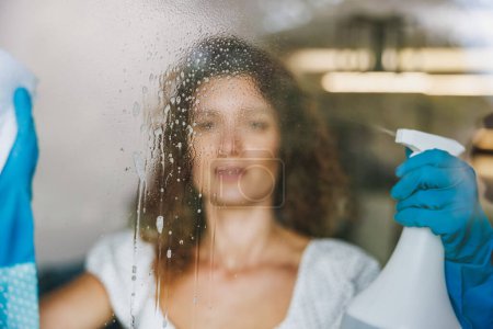 Téléchargez les photos : Femme nettoyage vitre avec un chiffon et pulvérisation pour l'hygiène des bactéries et de la poussière. Services d'entretien ménager ou d'accueil. - en image libre de droit
