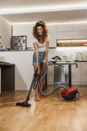 A smiling woman housekeeping cleaning a home. She is using a vacuum to clean a hard wood floor in a living room.
