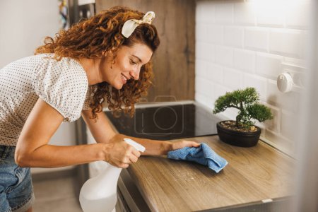 Téléchargez les photos : Jeune femme souriante faisant ses tâches quotidiennes à la maison. Elle nettoie et désinfecte un comptoir de cuisine.. - en image libre de droit