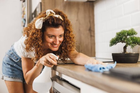 Téléchargez les photos : Jeune femme souriante faisant ses tâches quotidiennes à la maison. Elle essuie et désinfecte un comptoir de cuisine avec un chiffon . - en image libre de droit