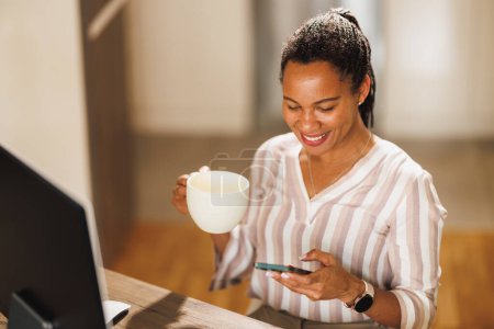 Photo for African business woman using a cellphone and drinking coffee while working from home. - Royalty Free Image