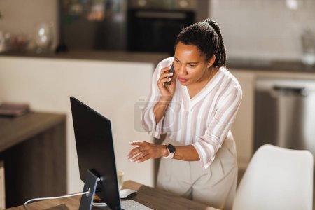 Téléchargez les photos : Femme d'affaires africaine utilisant un téléphone portable tout en travaillant sur un ordinateur à partir de son bureau à domicile. - en image libre de droit