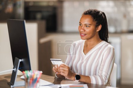 Photo for African business woman using her credit card while working on a computer at home. - Royalty Free Image
