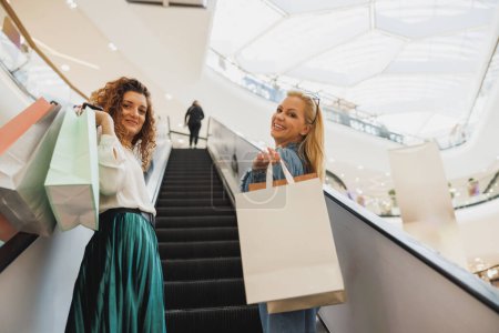 Téléchargez les photos : Tourné de deux jolies femmes debout sur l'escalator dans le centre commercial et souriant à la caméra pendant les ventes Black Friday ou Cyber Monday. - en image libre de droit