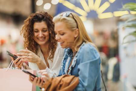 Téléchargez les photos : Plan recadré de deux belles femmes gaies surfant sur un smartphone tout en profitant d'une journée de shopping dans un centre commercial de la ville. - en image libre de droit