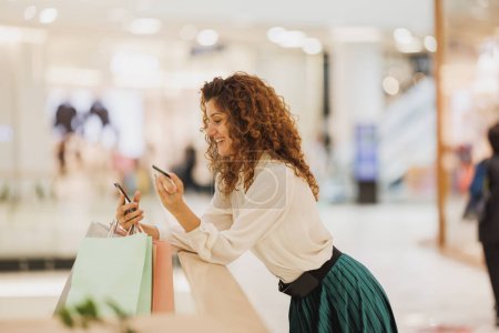 Téléchargez les photos : Prise de vue d'une femme souriante utilisant son smartphone pour vérifier les offres de vente en ligne ou en utilisant une application de réduction lors du magasinage dans un centre commercial. - en image libre de droit