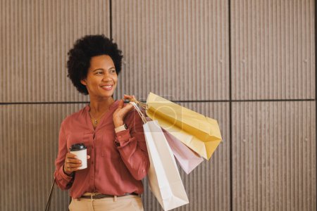 Téléchargez les photos : Prise de vue d'une femme noire portant des sacs à provisions et tenant une tasse de café à emporter debout contre un mur dans un centre commercial. - en image libre de droit
