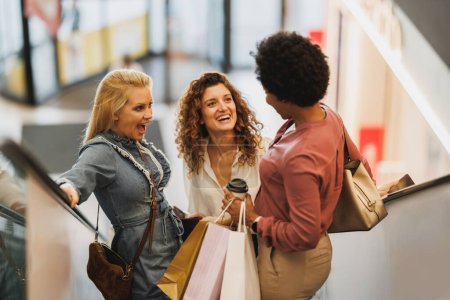 Téléchargez les photos : Tourné de trois femmes souriantes qui s'amusent et se tiennent debout sur l'escalator dans un centre commercial ensemble lors d'une virée shopping. - en image libre de droit