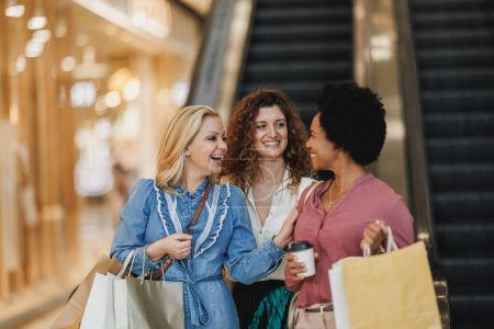 Téléchargez les photos : Tourné de trois femmes souriantes parlant et prenant un peu de temps pour profiter d'une virée shopping dans le centre commercial de la ville. - en image libre de droit