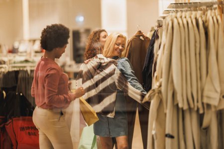 Foto de Fotografía de una mujer atractiva sosteniendo una prenda mientras pide consejo a sus amigos en una tienda de ropa. - Imagen libre de derechos