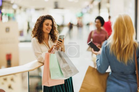 Téléchargez les photos : Femme élégante utilisant un téléphone portable tout en portant des sacs lors d'une virée shopping au centre commercial. - en image libre de droit