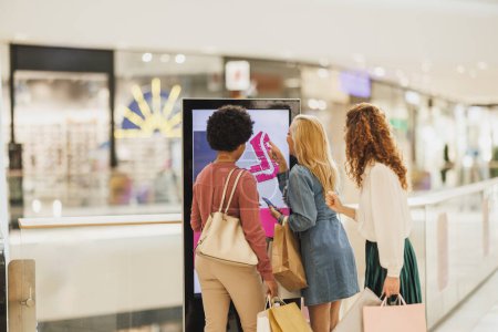 Téléchargez les photos : Vue arrière d'un heureux trois amies à la recherche d'un magasin sur une carte au centre commercial. - en image libre de droit