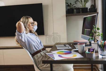 Téléchargez les photos : Jeune femme créative assise avec les mains derrière la tête et relaxante tout en travaillant dans le confort de sa maison. - en image libre de droit