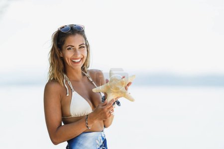 Foto de Hermosa mujer joven sosteniendo estrellas de mar y disfrutando en la playa. Mirando la cámara. - Imagen libre de derechos