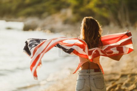 Téléchargez les photos : Vue arrière d'une femme avec drapeau national américain profitant d'une journée de détente sur la plage. - en image libre de droit