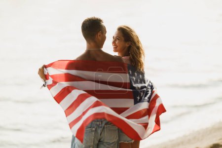 Téléchargez les photos : Vue arrière d'un couple aimant avec drapeau national américain profitant d'une journée de détente sur la plage. - en image libre de droit