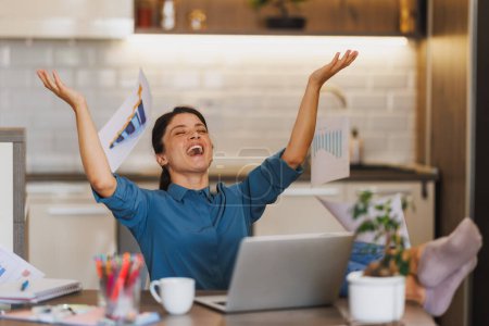 Photo for Young female entrepreneur celebrating with raised up her hands in surprise and throwing papers in the air  as she works from her home office. - Royalty Free Image