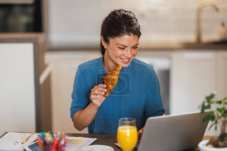 Photo for Business woman eating a croissant for breakfast while working on a laptop at home. - Royalty Free Image