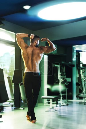 Foto de Vista trasera de un culturista muscular que muestra sus músculos perfectos mientras posa después de hacer ejercicio en el gimnasio. - Imagen libre de derechos
