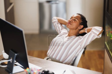 Téléchargez les photos : Femme d'affaires africaine assis avec ses mains derrière la tête et relaxant tout en travaillant à partir de son bureau à domicile. - en image libre de droit