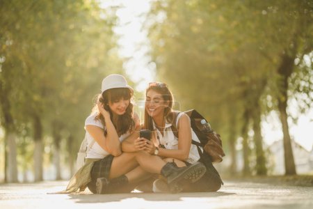Photo for Two cute smiling young women are surfing social media during walking along the sunny avenue. They are looking at the photos on a smartphone. - Royalty Free Image