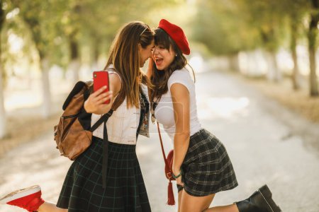 Two beautiful cheerful girl friends are making selfie photos during walking along the sunny avenue.