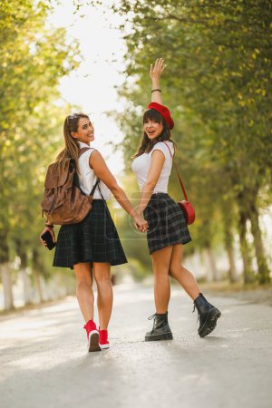 Rückansicht von zwei gut gelaunten jungen Frauen, die die sonnige Herbstallee entlanggehen und in die Kamera schauen.