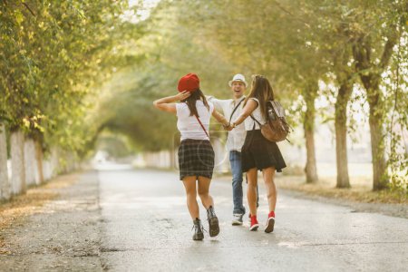 Three beautiful cheerful friends are having fun while walking along the sunny avenue and holding hands.