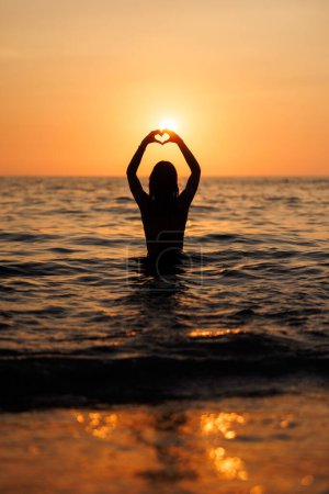 Téléchargez les photos : Silhouette d'une jeune femme profitant du coucher de soleil sur la plage. La lumière du soleil brille à travers ses mains faisant forme de coeur. - en image libre de droit