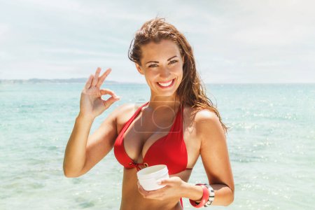 Photo for Portrait of a beautiful young woman applying sunscreen cream on the beach. She is smiling and looking at camera. - Royalty Free Image