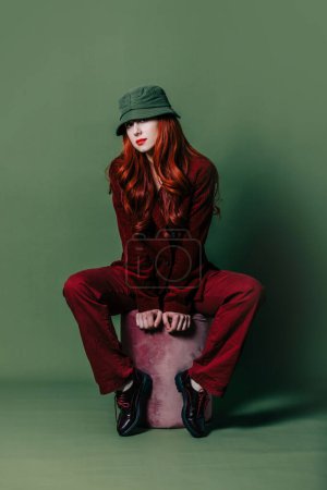 Téléchargez les photos : Stylish woman in sunglasses, hat and red suit sits on green background - en image libre de droit