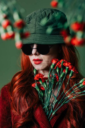 Foto de Stylish redhead woman in hat and burgundy color suit with carnations on green background - Imagen libre de derechos