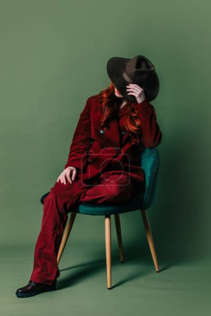 Photo for Stylish woman in hat and retro red suit sits in chair on green background - Royalty Free Image
