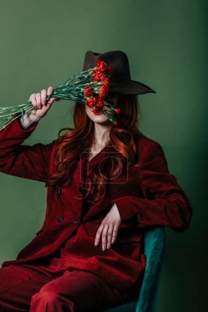 Foto de Stylish redhead woman in hat and burgundy color suit on green background - Imagen libre de derechos