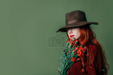 Téléchargez les photos : Stylish redhead woman in hat and burgundy color suit with carnations on green background - en image libre de droit
