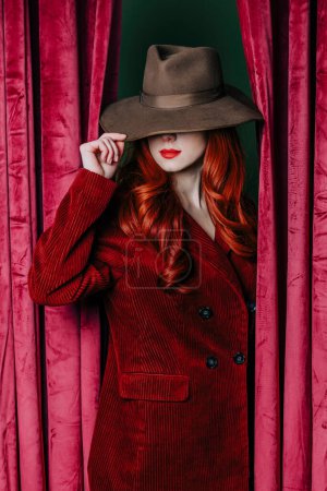 Foto de Stylish redhead woman in hat peek through the viva magenta color curtains - Imagen libre de derechos