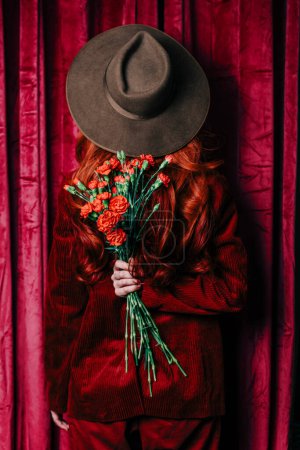 Téléchargez les photos : Stylish redhead woman in hat with carnations peek through the viva magenta color curtains - en image libre de droit