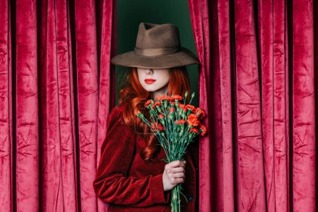 Foto de Stylish redhead woman in hat with carnations peek through the viva magenta color curtains - Imagen libre de derechos