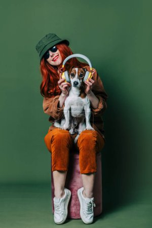 Foto de Stylish redhead woman with headphones and dog on green background - Imagen libre de derechos