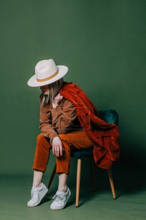 Téléchargez les photos : Stylish blond hair woman in hat nad orange pants sits in chair on green backgorund in studio - en image libre de droit
