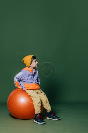 Photo for Stylish little boy in headphones sits on orange swiss ball on green background - Royalty Free Image
