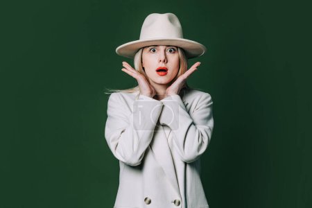Foto de Suprised stylish blond hair woman in white blazer and hat on green background - Imagen libre de derechos
