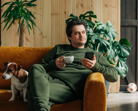 Téléchargez les photos : Caucasian man sits in sofa with coffee cup and smartphone at home - en image libre de droit