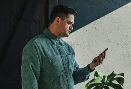 Foto de Caucasian guy in green shirt looking on display of smartphone at home, whadow on wall on background - Imagen libre de derechos