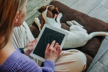 Foto de Mujer de pelo rubio se sienta en el suelo con la tableta y un perro. Vista lateral trasera - Imagen libre de derechos