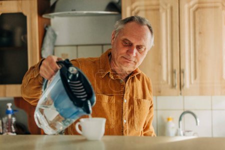 Foto de Elderly man pours water into a cup from the filter in the kitchen - Imagen libre de derechos