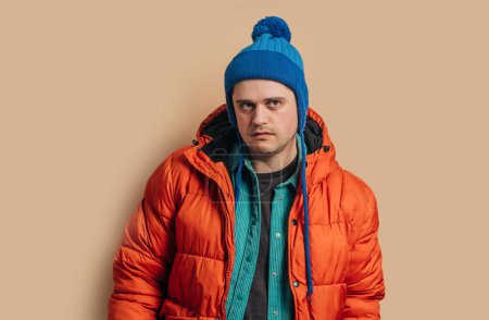 Photo pour Funny caucasian guy in blue hat, orange down jacket on brown background - image libre de droit