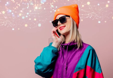 Téléchargez les photos : Style fille dans les années 80 costume de sport et lunettes de soleil tenir téléphone mobile sur fond rose - en image libre de droit