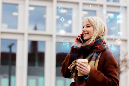 Photo for Beautiful blonde woman with cup of coffee talking by mobile phone on city street - Royalty Free Image