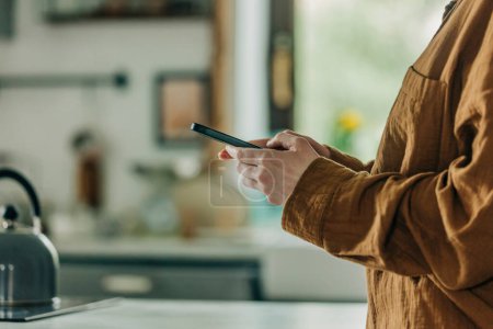 Foto de Mujer con camisa marrón sostiene el teléfono inteligente en las manos en la cocina y mirando hacia adentro para mostrar. Vista lateral con cocina en bokeh sobre fondo - Imagen libre de derechos